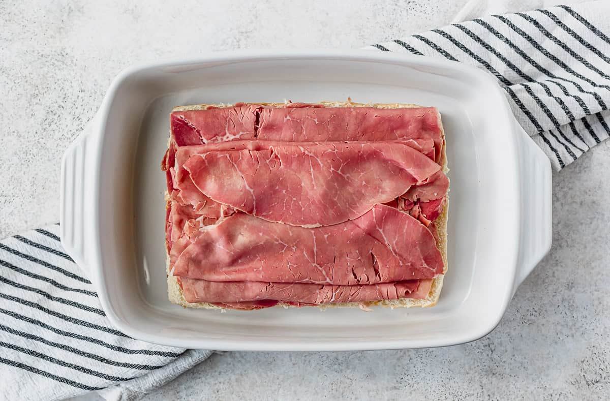Corned beef layered on top of rolls in a white casserole dish over a striped towel and gray background.