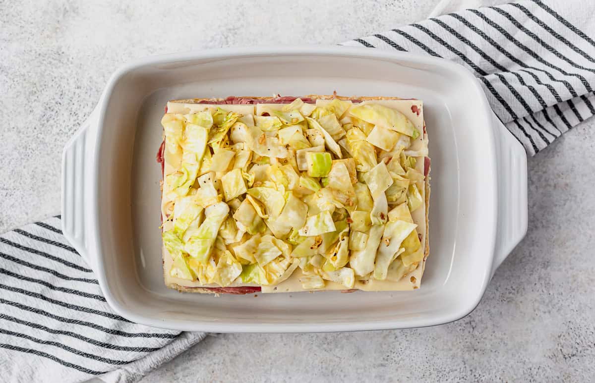 Chopped, cooked cabbage on top of corned beef and Swiss cheese in a white casserole dish over a striped towel and gray background.