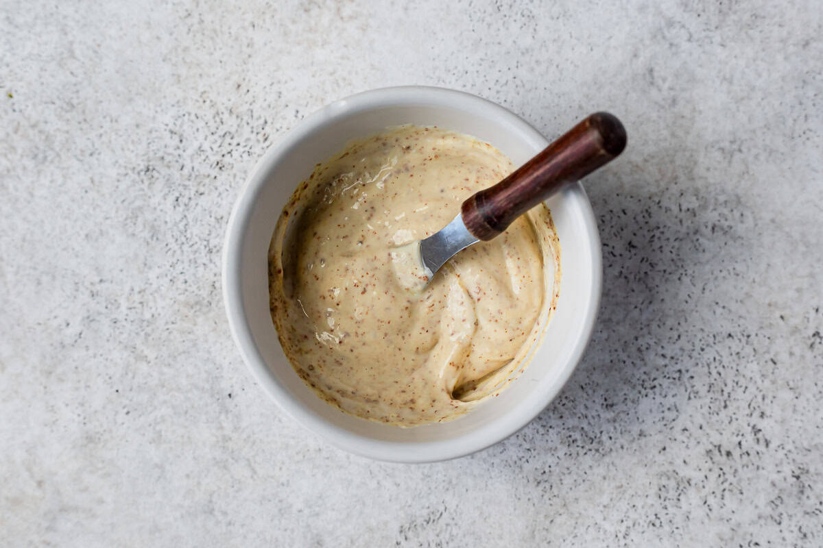 Mixed mayonnaise and dijon mustard in a white bowl with a wood handled spreader in it.
