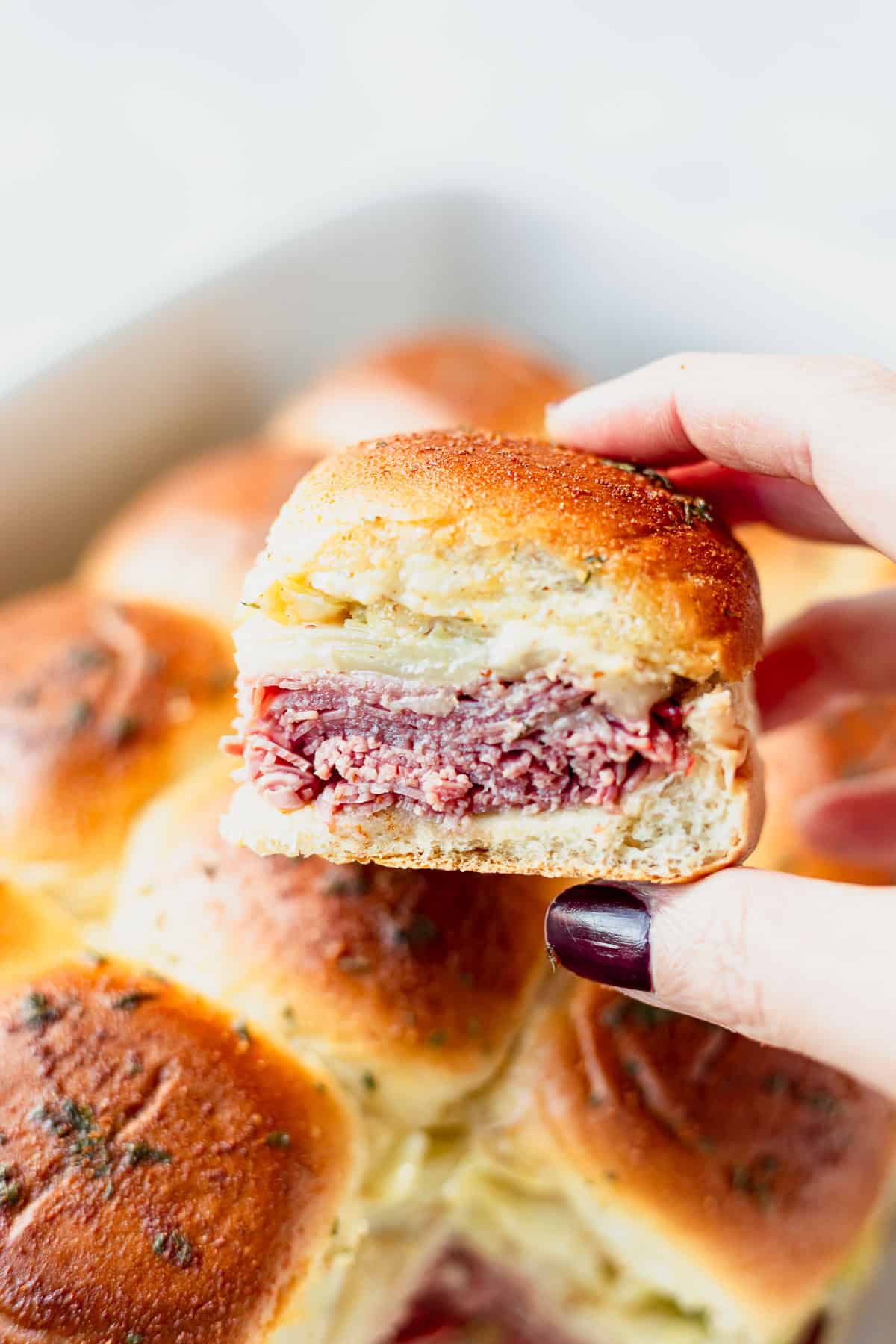 A hand holding a corned beef slider over the casserole dish filled with more sliders.