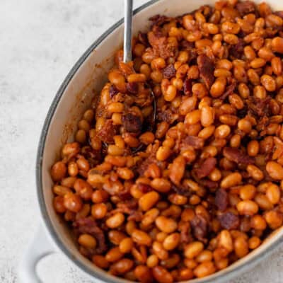 Baked beans in an oval baking dish with a spoon in them over a light background.