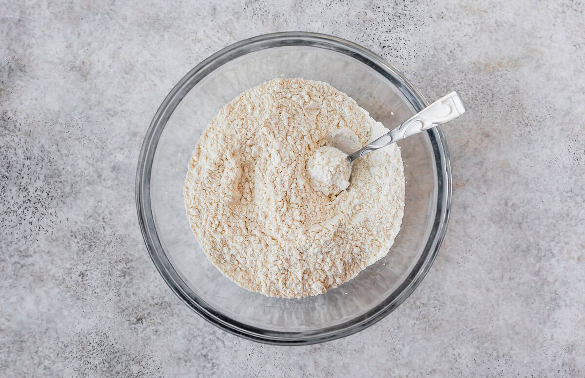 Dry ingredients for biscuits in a glass bowl with a spoon over a gray background.