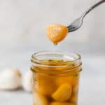 A garlic clove with oil dripping off of it being held over a jar of garlic confit.