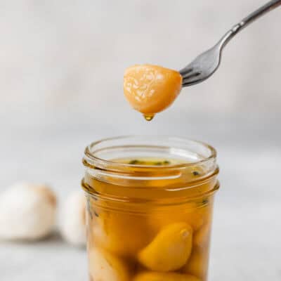 A garlic clove with oil dripping off of it being held over a jar of garlic confit.