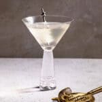 A Gibson Cocktail in a martini glass with a gold cocktail strainer in the foreground.