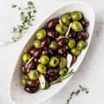 An oval white bowl filled with green and purple olives with sprigs of thyme around it.