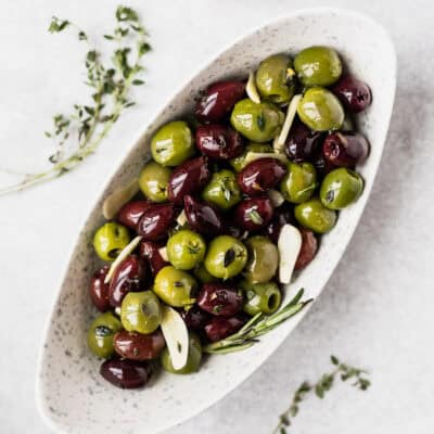 An oval white bowl filled with green and purple olives with sprigs of thyme around it.
