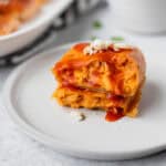 A buffalo chicken egg roll cut in half and stack on each other on a white plate with buffalo sauce running down it and blue cheese on top.