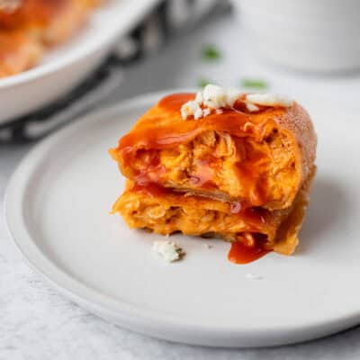 A buffalo chicken egg roll cut in half and stack on each other on a white plate with buffalo sauce running down it and blue cheese on top.