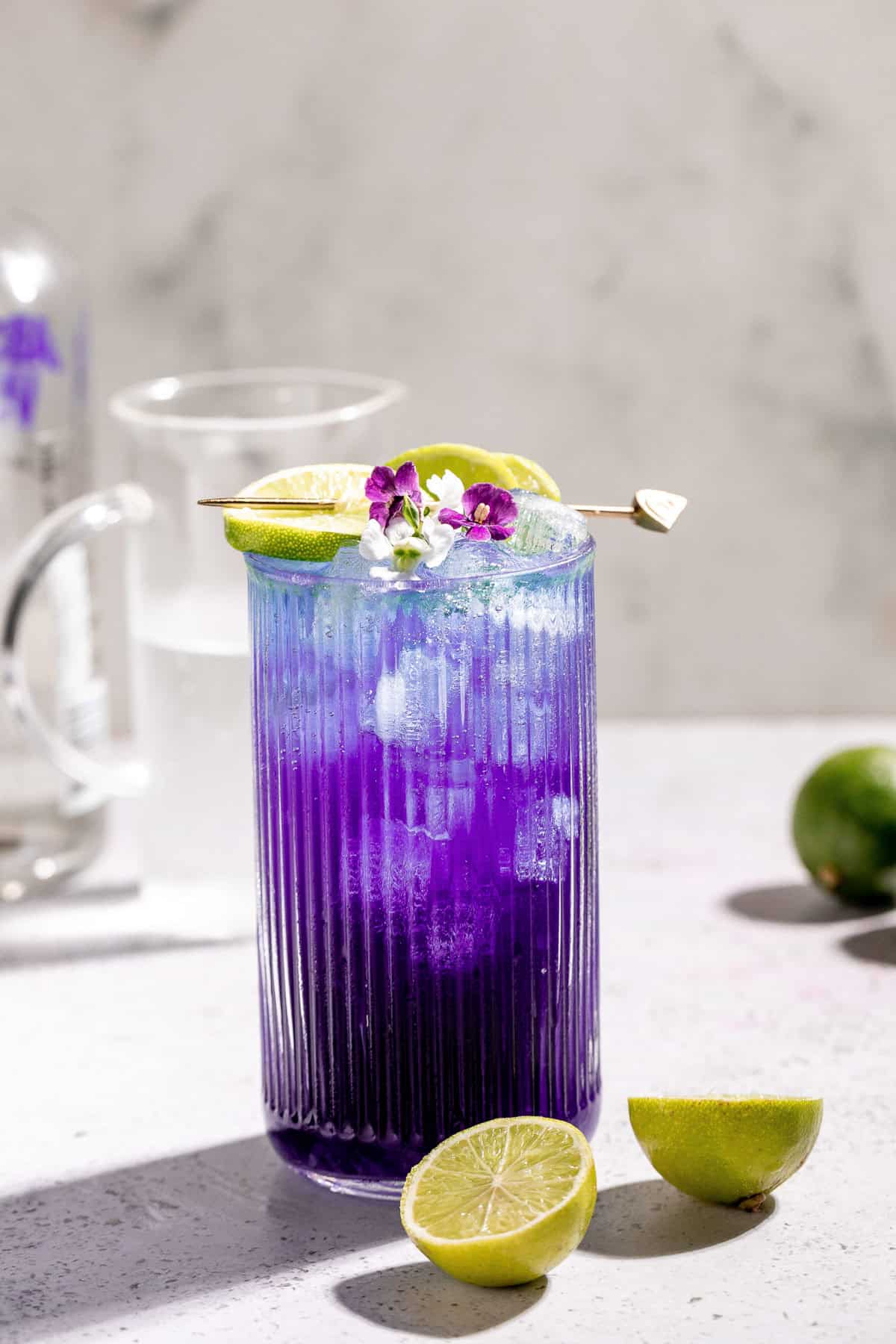A purple rain cocktail garnished with lime wedges and purple flowers on a white surface with limes around it and bottles in the background.