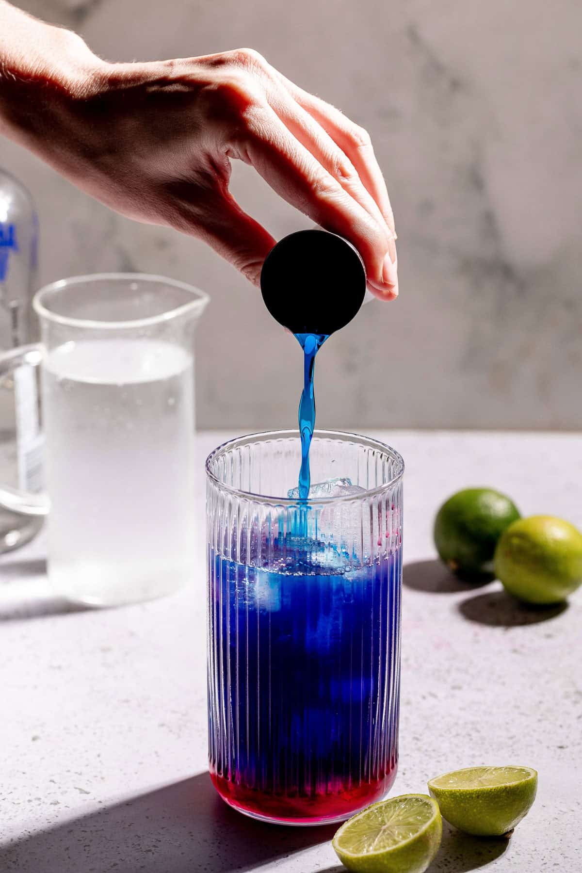 Ingredients being poured into a tall glass with limes and a clear pitcher in the background.