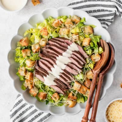 Overhead view of a steak caesar salad in a large scalloped bowl with wood serving tongs on the side, over a striped linen with bowls of ingredients around.