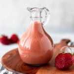 A cruet filled with strawberry balsamic vinaigrette on a wood board with strawberries around it.