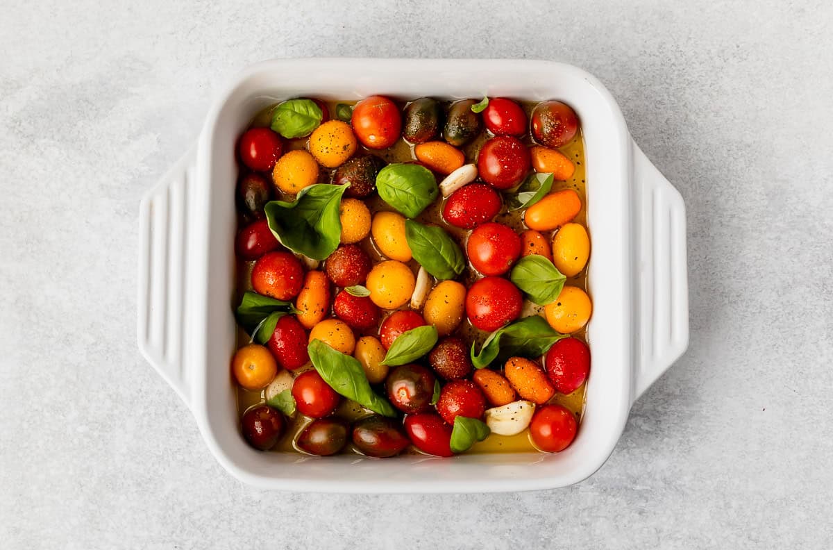 Tomatoes, garlic and basil in oil in a white, square baking dish before roasting.