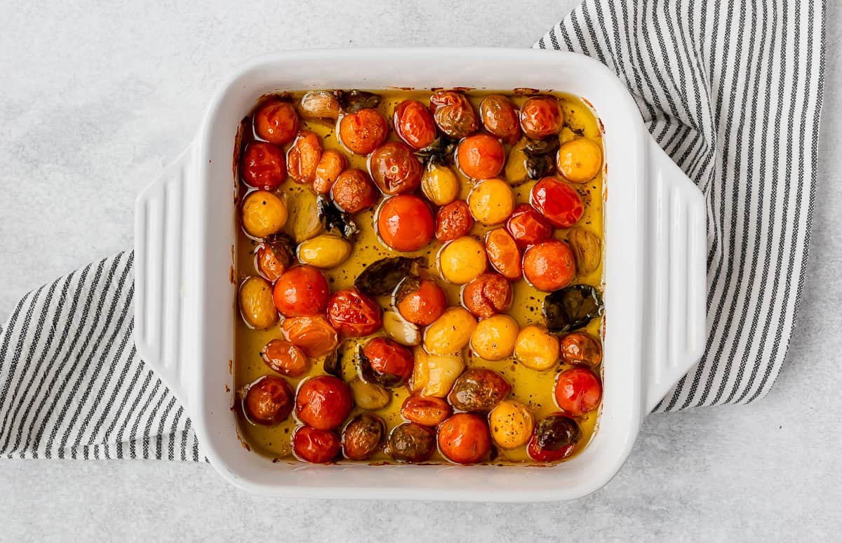 Roasted cherry tomato confit with garlic and basil in oil in a white, square baking dish.