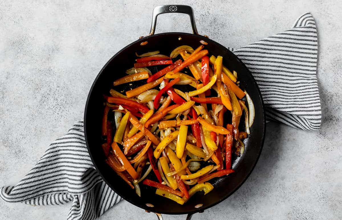 Strips of peppers and onions with garlic in a black skillet on a striped towel over a light background.