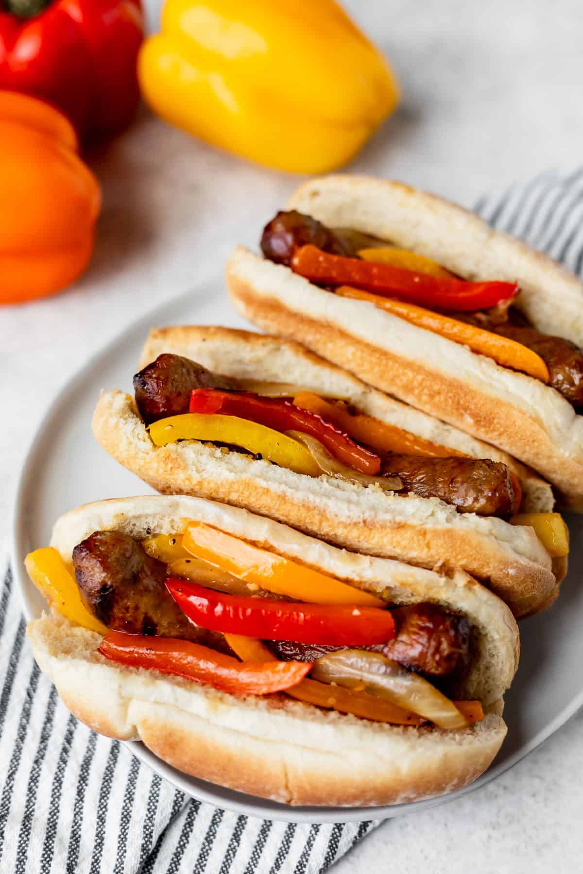 Three grilled sausage and peppers sandwiches on a plate over a striped towel with bell peppers in the background.