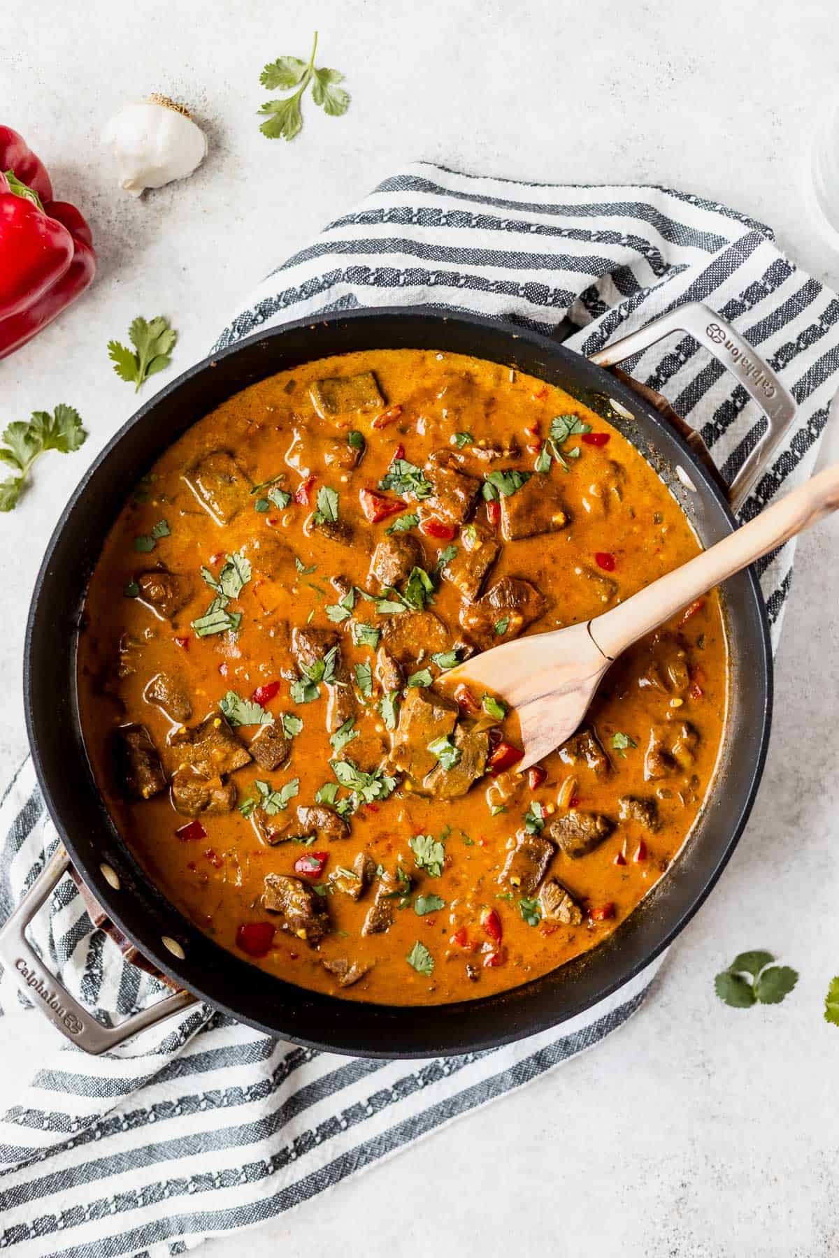 Overhead view of a black skillet filled with coconut curry beef and sauce over. striped linen with a pepper, garlic and cilantro around it.