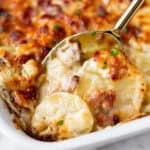A spoon pushing up some scalloped potatoes and ham in a white casserole dish.