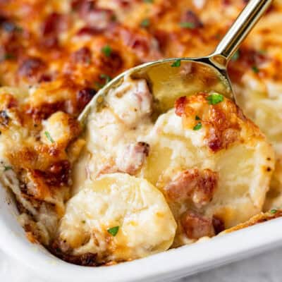 A spoon pushing up some scalloped potatoes and ham in a white casserole dish.