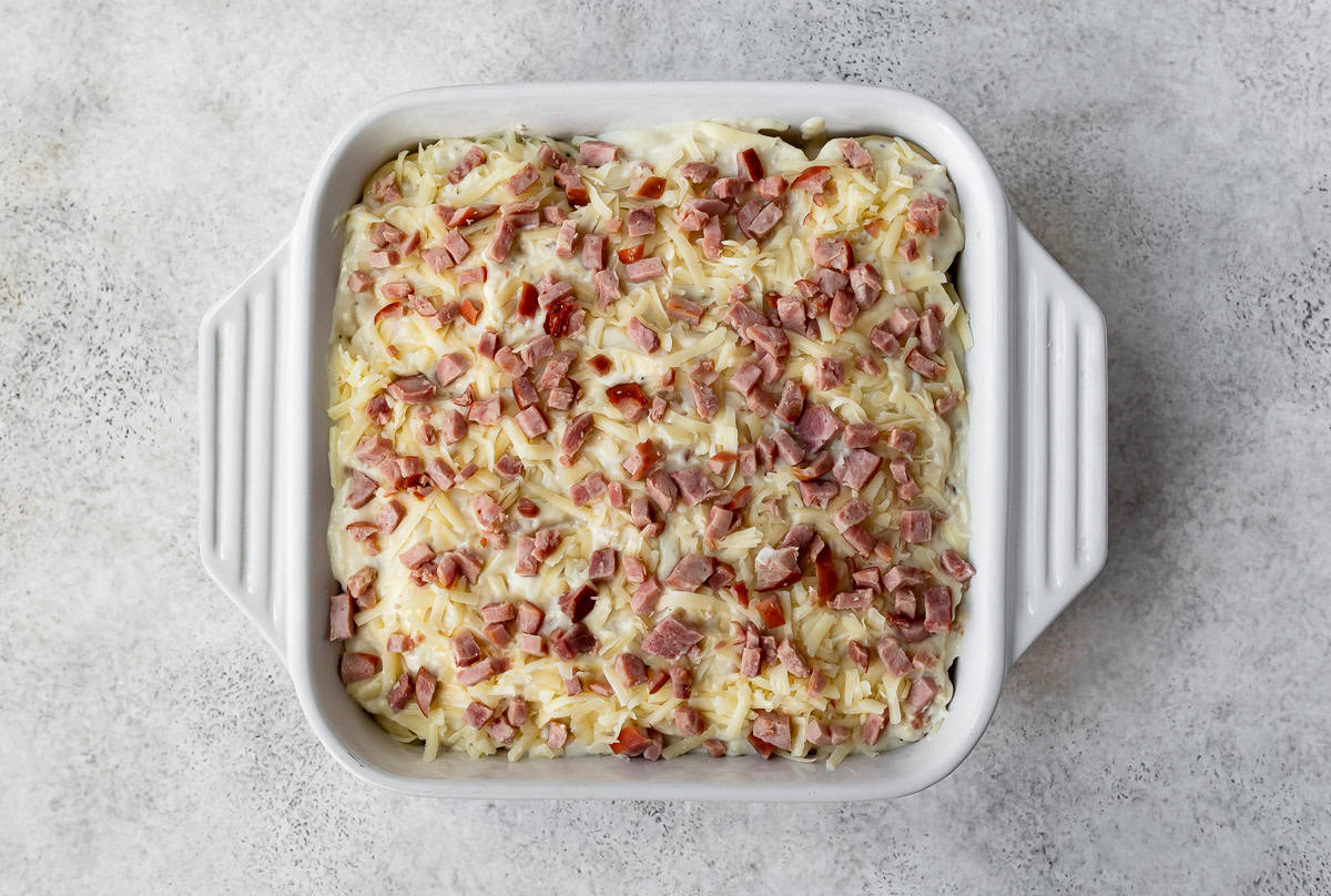 Layers of potatoes with cheese sauce and ham over the tops in a white, square baking dish.