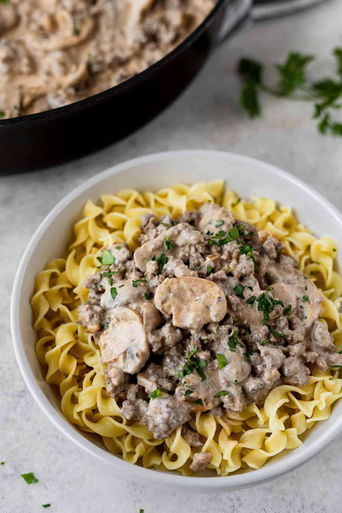 A white bowl filled with egg noodles topped with ground beef stroganoff with the skillet in the background.