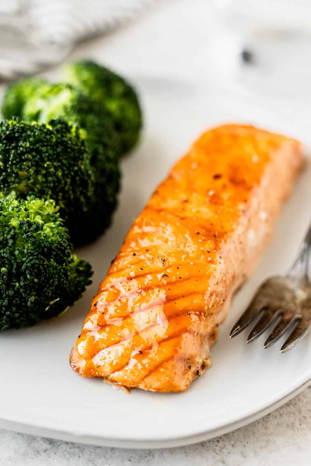 A filet of maple brown sugar glazed salmon and broccoli on a white plate with a fork.