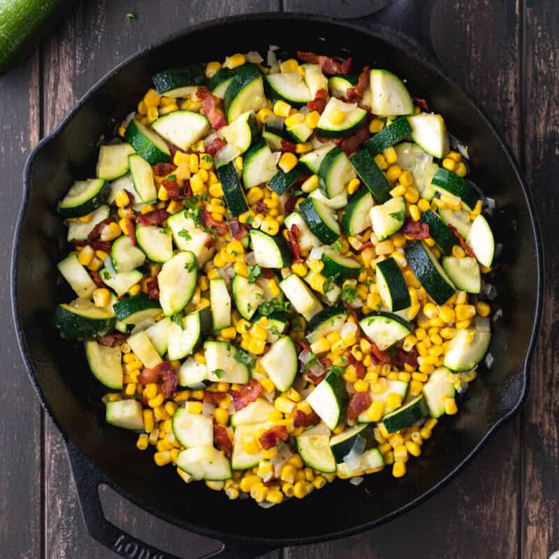 Overhead view of fried zucchini corn and bacon skillet over a wood background.