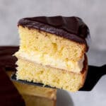 A slice of Boston Cream Pie on a pie server being lifted up from the rest of the cake.