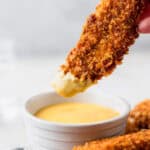 A corn flake coated chicken tender with honey mustard on the end being help up over the bowl of dipping sauce.