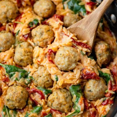 Close up of Tuscan orzo with sun-dried tomatoes and spinach topped with baked chicken meatballs with a wood spoon in it.