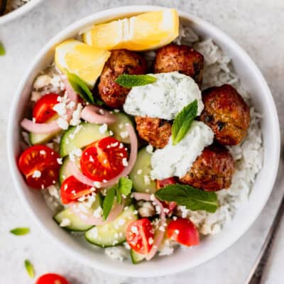 A white bowl with rice, Greek chicken meatballs, tomatoes, cucumber, onions, lemons and tzatziki.