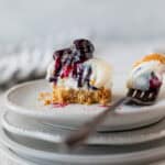 A mini blueberry cheesecake on a stack of small plates with a bite taken out on a fork next to it on the plate. There is a striped linen around it.