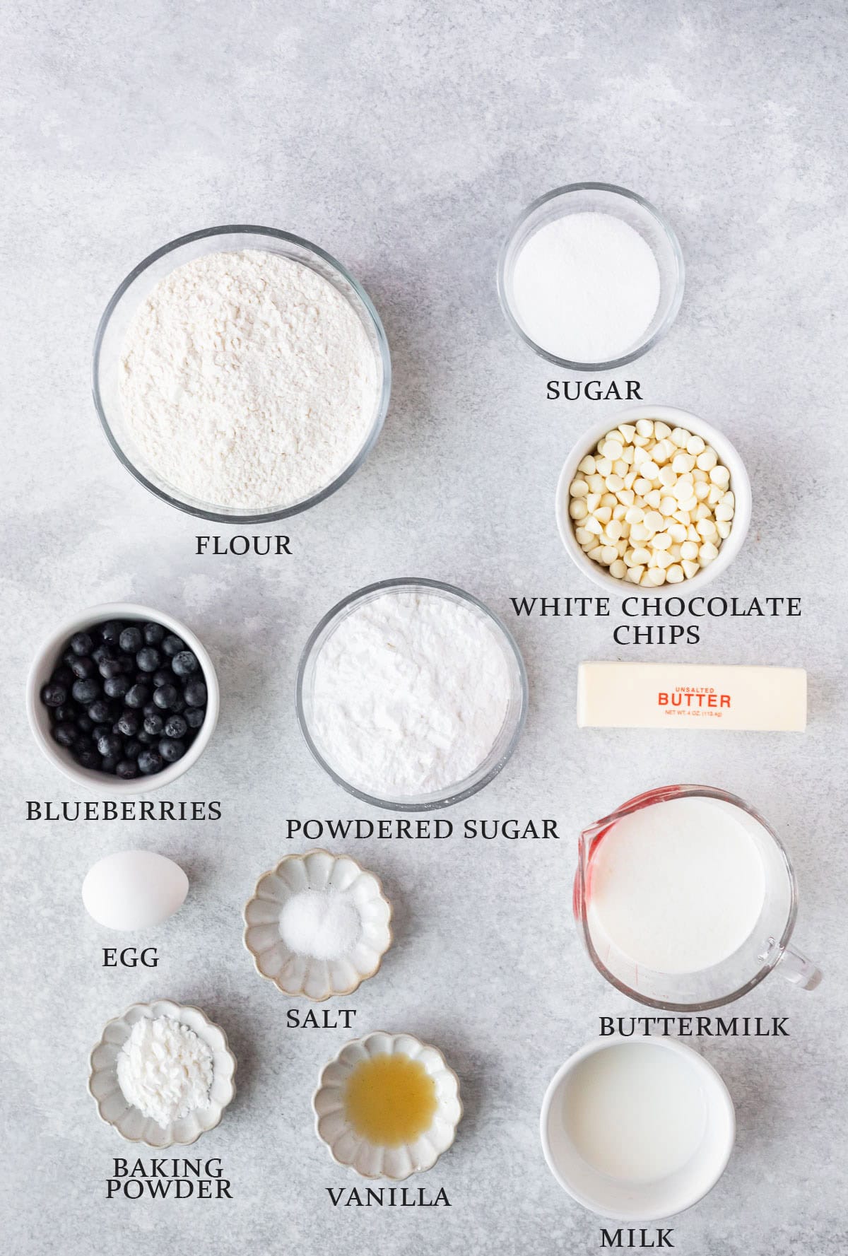 Ingredients needed to make white chocolate blueberry scones on a light surface with text overlay that reads "flour, sugar, egg, blueberries, vanilla, baking powder, salt, buttermilk, milk, white chocolate chips, butter and powdered sugar."