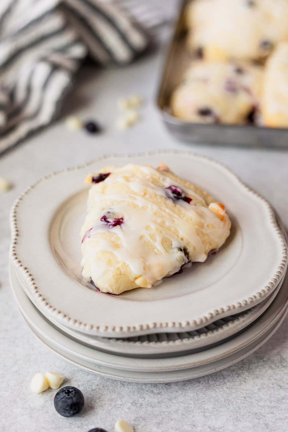 A single white chocolate chip blueberry scone on a stack of 4 small white plates with a tray of scones and striped linen in the background.