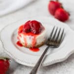 A mini no-bake strawberry cheesecake on a small plate with a fork on it and strawberries around it.