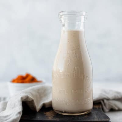 Homemade pumpkin spice coffee creamer in a glass jar on a wood board with a white bowl of pumpkin puree and a tan linen in the background.