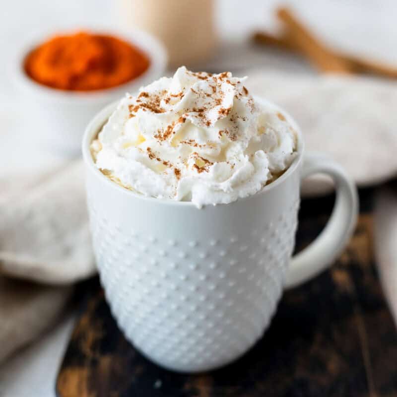 A pumpkin spice latte topped with whipped cream and pumpkin spice in a white mug over a wood board with a bowl of pumpkin puree, cinnamon sticks and jar of coffee creamer in the background.