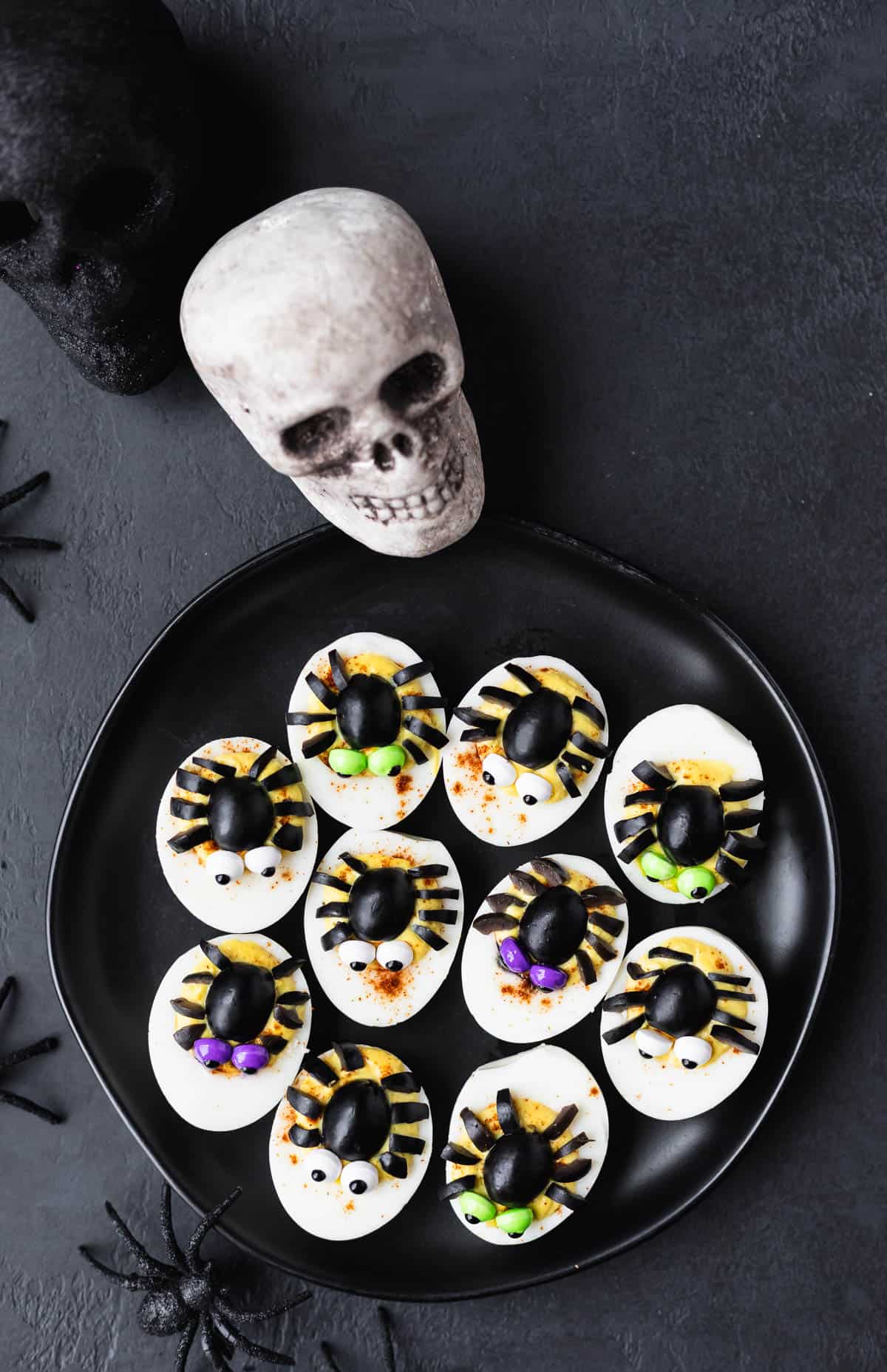 Overhead view of 10 spider deviled eggs on a black plate over a black background with black spiders and black and gray skulls around the plate.